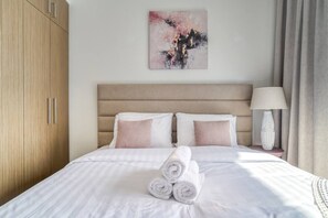 A neatly made bed with rolled towels, a headboard, a bedside lamp, and a painting on the wall.