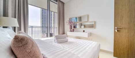 A modern hotel room with a large bed, a bedside lamp, a wooden door, a balcony with a view, and a small table with a chair.