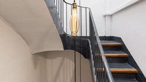 Una escalera con barandas negras y escalones de madera, que conduce a un rellano con un muro curvo y lámparas colgantes.