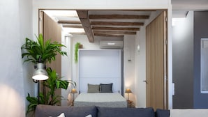 Un dormitorio compacto con cama, lámparas de noche, una planta en maceta y vistas al área de estar contigua.
