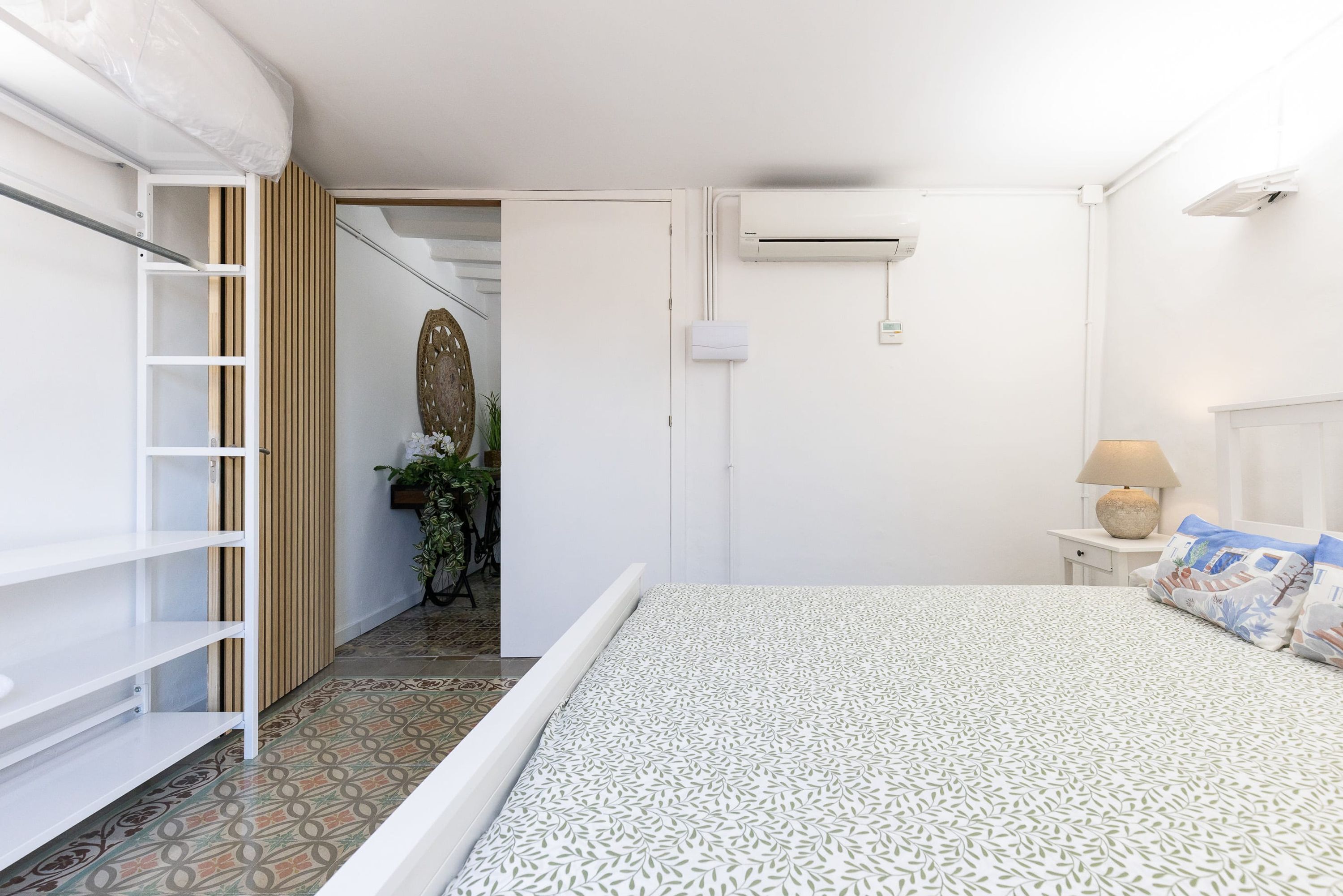 Un dormitorio con cama, mesita de noche, lámpara, aire acondicionado instalado en la pared y puerta que da a otra habitación.