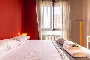 Una habitación de hotel con cama, una ventana con cortinas, una lámpara de noche y una pila de toallas.