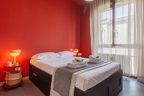 Ein Hotelzimmer mit Bett, Nachttisch mit Lampe, Fenster mit Vorhängen und Heizkörper.
