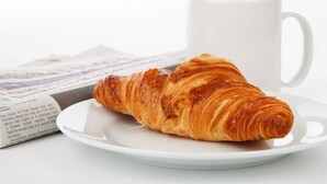 Een croissant op een wit bord, met een witte mok op de achtergrond.