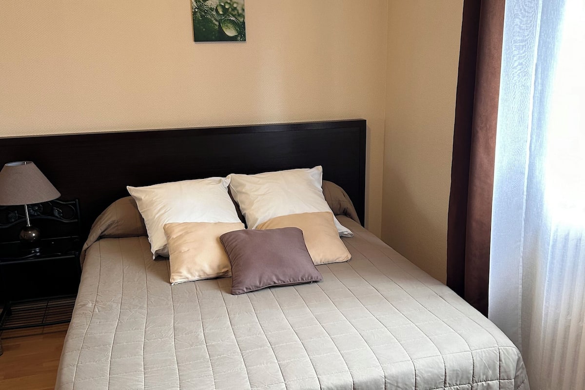Una cama bien hecha con colcha acolchada y almohadas, una mesita de noche con lámpara, y una ventana con cortinas.
