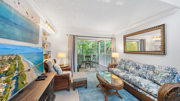 A spacious living room with a large window, a sofa, a coffee table, and a painting of a beach scene. - Beach Castle Resort, 2nd Floor, Updated, Walk to Beach/Bay, Pool (Longboat Key)