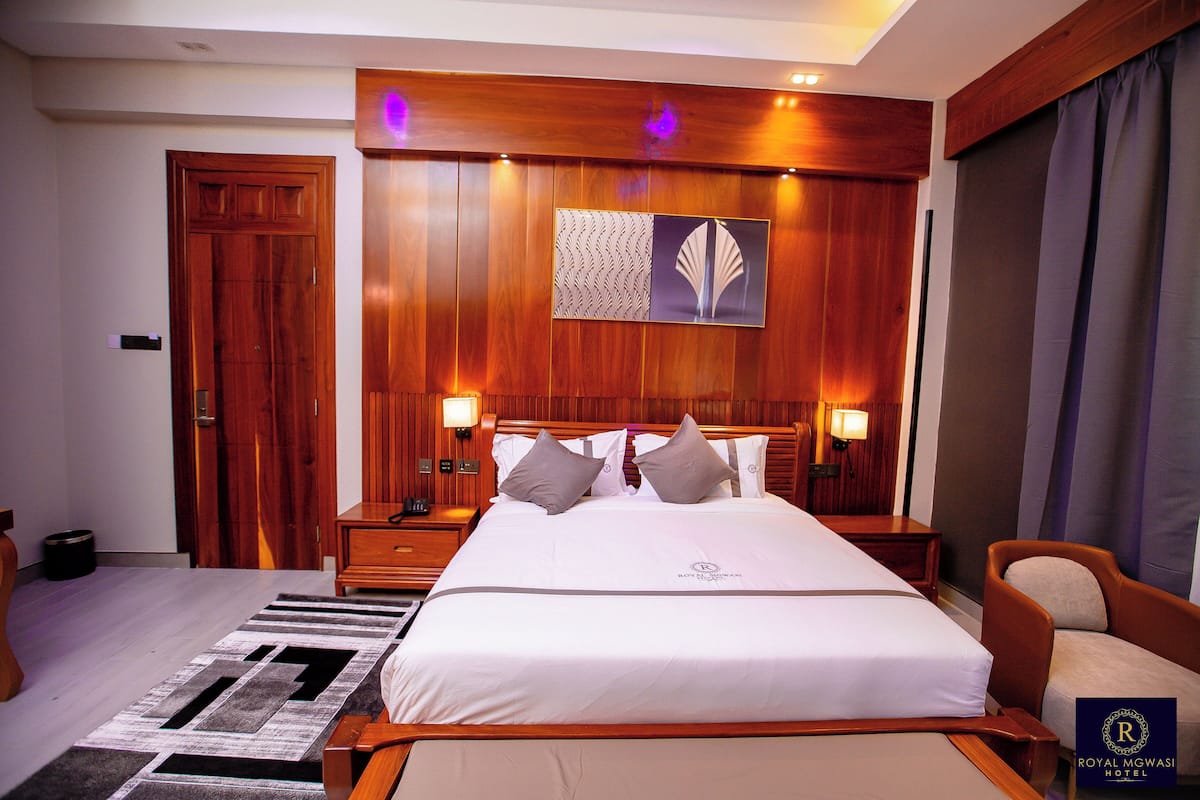 Quarto de hotel com uma cama grande, cabeceira de madeira, duas mesas de cabeceira com luminárias, um criado-mudo, uma cadeira e uma obra de arte na parede.