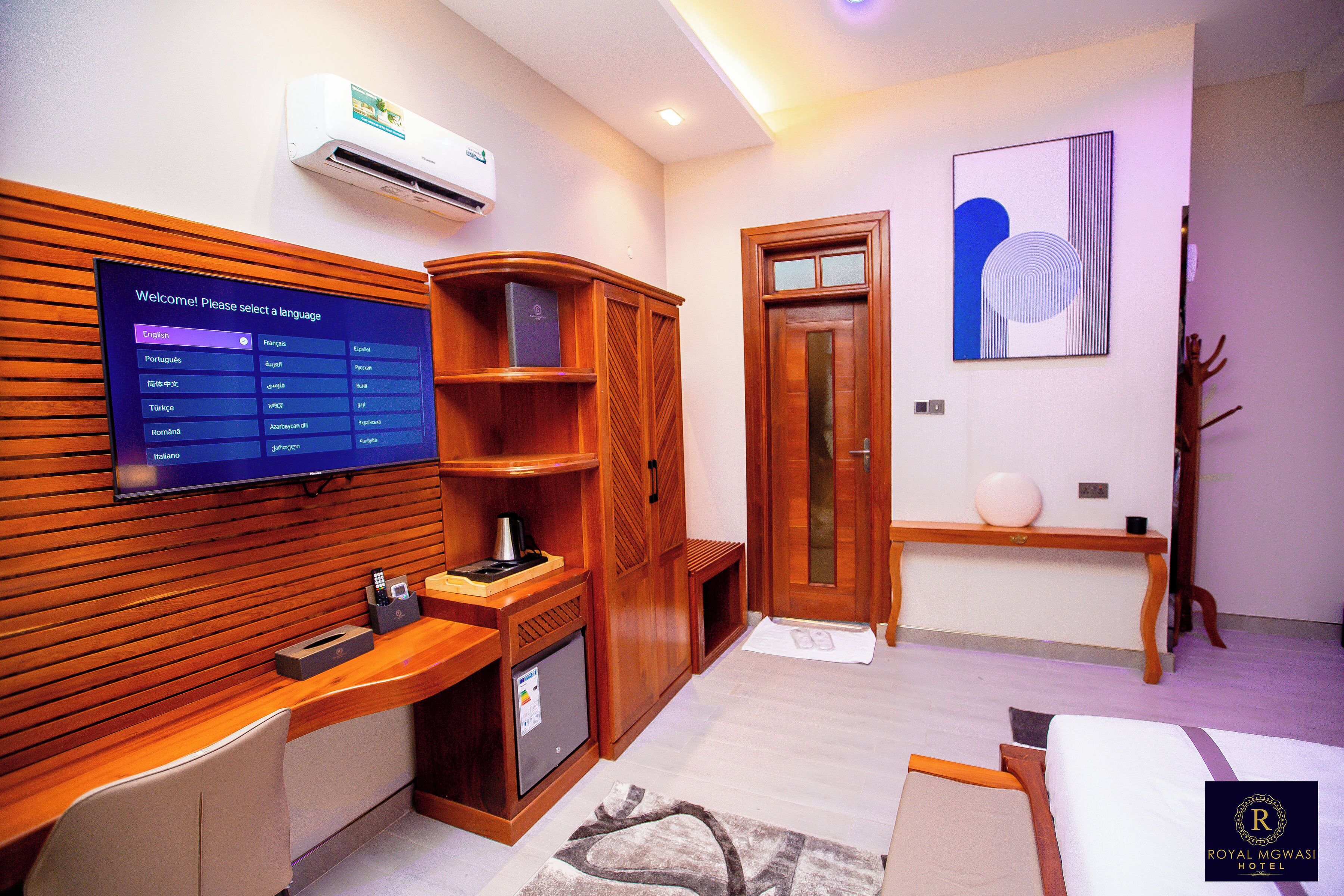 A hotel room with a wooden reception desk, a television displaying a language selection menu, a small refrigerator, a wooden wardrobe, a door with a glass panel, and a wall-mounted artwork.