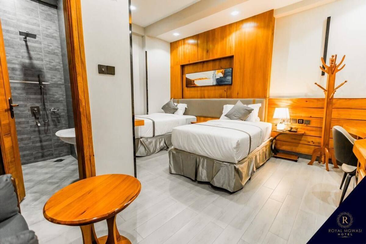 Quarto de hotel com duas camas, uma mesa de madeira, chuveiro e um cabideiro.