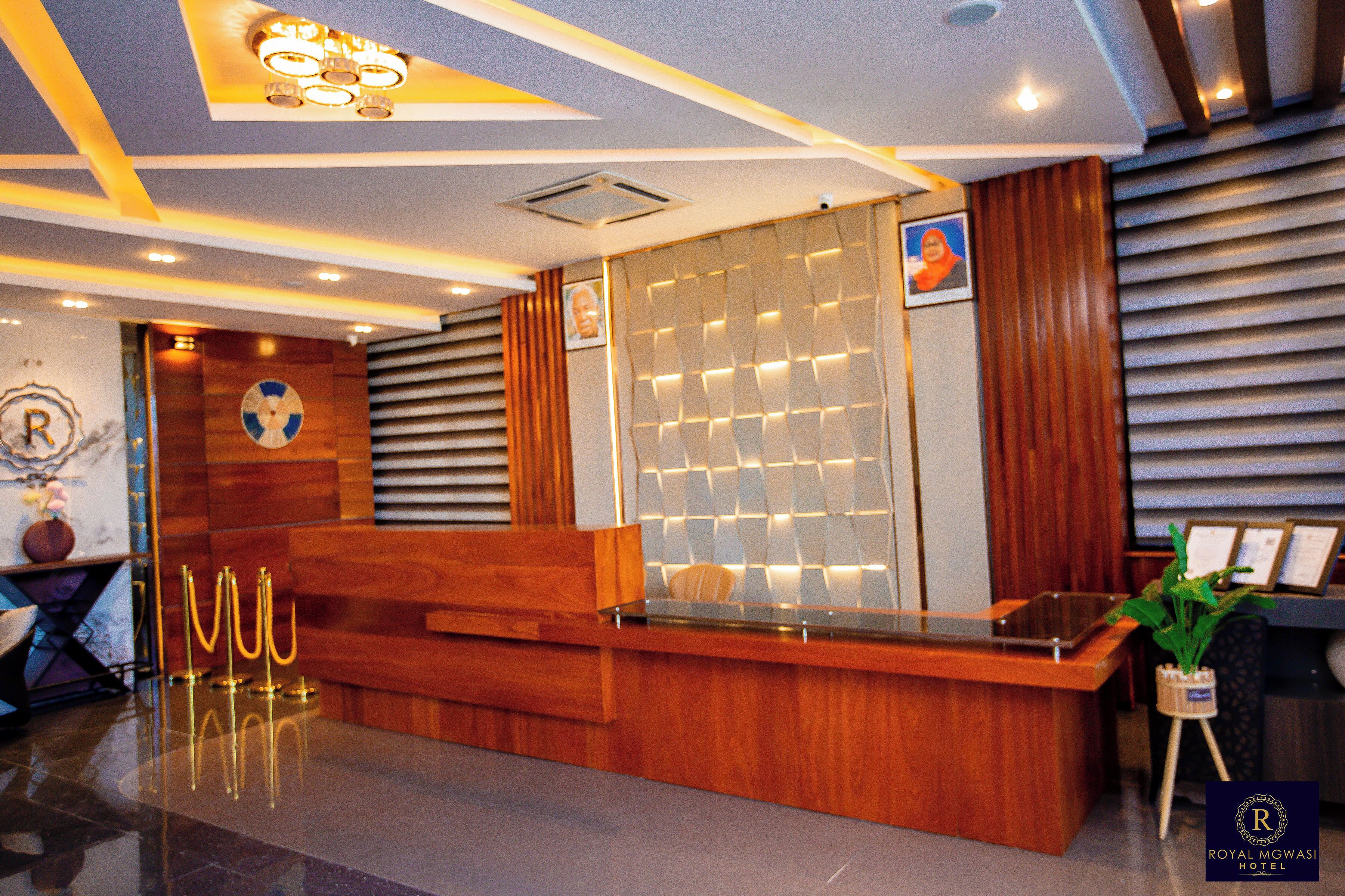 A hotel reception area with a wooden reception desk, a wall-mounted photo, and a potted plant.