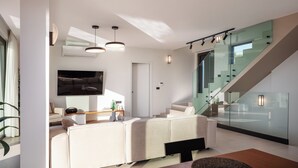 A modern living room with a sofa, a flat-screen TV, and a staircase with glass railings.