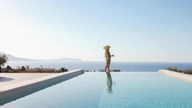 A person standing at the edge of a infinity pool with a view of the sea and mountains in the background.
