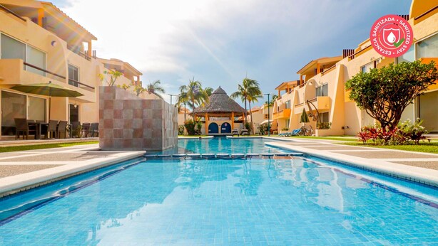 Una piscina con un gazebo dal tetto di paglia al centro, circondata da palme e poltrone prendisole.