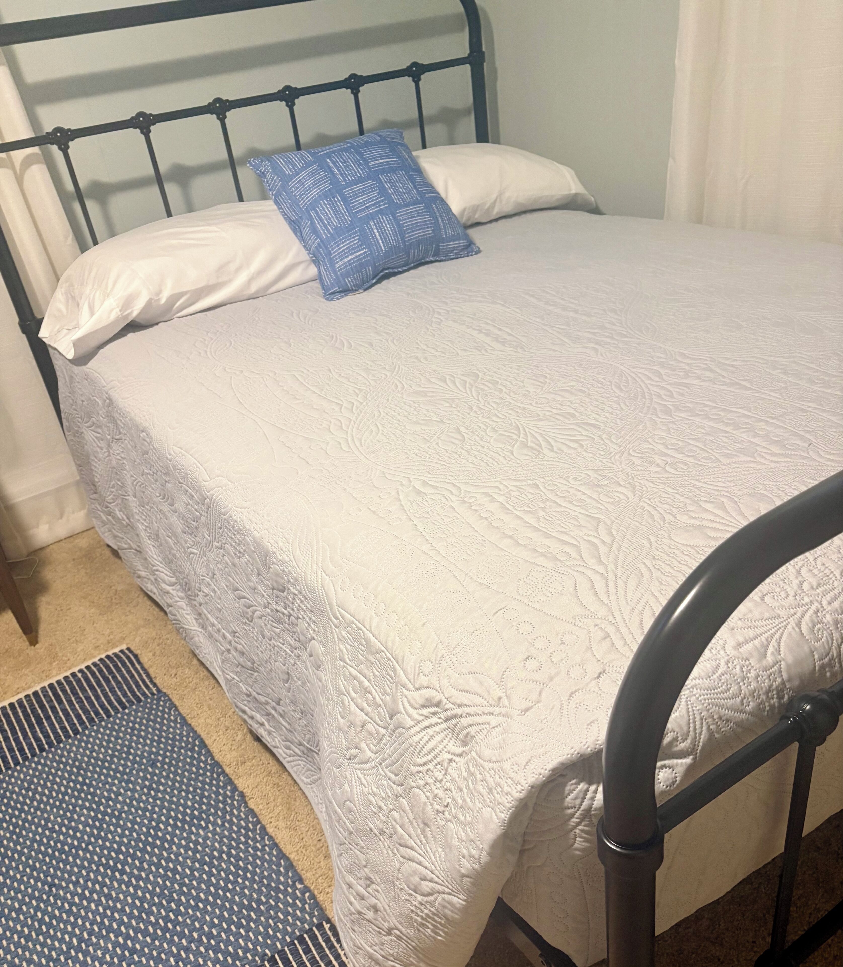 Una cama con colcha blanca, almohada azul, cabecera de metal y una alfombra a rayas en el suelo.
