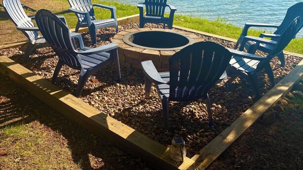 Ein Seeufer mit einer Holzterrasse, Adirondack-Stühlen, einer Feuerstelle und einem Steg, der ins Wasser führt.