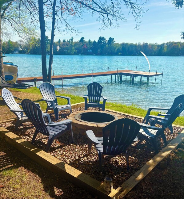 A lakeside deck with Adirondack chairs, a fire pit, and a dock extending into the water.