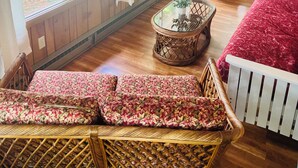 Ein gemütliches Zimmer mit Holzbalkendecke, großen Fenstern mit weißen Vorhängen, einem Korbssessel und einer roten gepolsterten Bank sowie einem kleinen runden Tisch mit einer Pflanze.