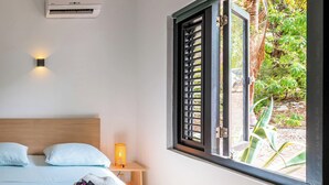 A bedroom with a bed, bedside lamp, and a window with a view of greenery.