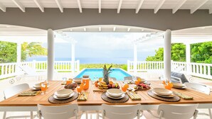 A covered dining area with a pool view, white furniture, and a table set with fruits and drinks.