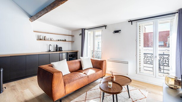 Un salon moderne avec un canapé en cuir marron, une petite table en bois et un coin cuisine doté de meubles sombres et d’un plan de travail blanc.