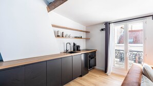 Une cuisine moderne avec des meubles de couleur sombre, un plan de travail en bois et un four encastré. Des étagères au-dessus du plan de travail servent à ranger divers ustensiles de cuisine. Une fenêtre donne sur un immeuble.