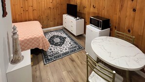 Een compacte kamer met houten paneelwanden, een bed met een geruit plaid, een witte kapstok, een klein wit tafeltje met een marmeren blad, een zwart-wit vloerkleed en een magnetron op een klein wit kastje.