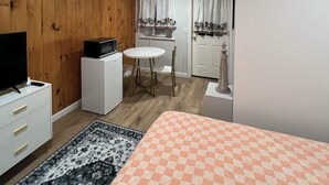 Een knus ingerichte kamer met houten wanden, een wit bureau, een kleine koelkast, een televisie, een ronde tafel met stoelen, een kamerbreed tapijt met dessin en een bed met een geruit sprei.