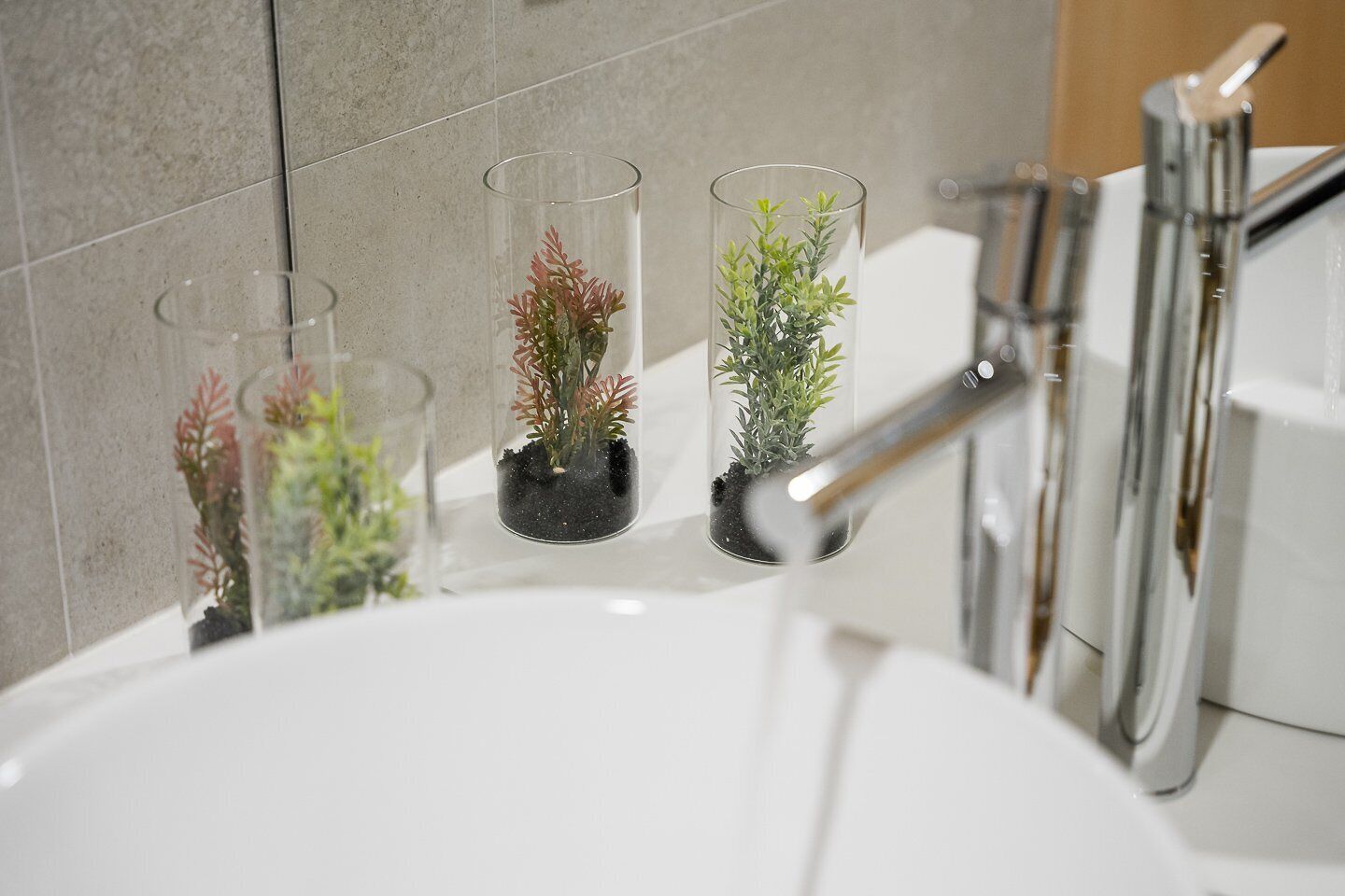 A bathroom with a white sink, three clear glass vases with plants, and a chrome faucet.