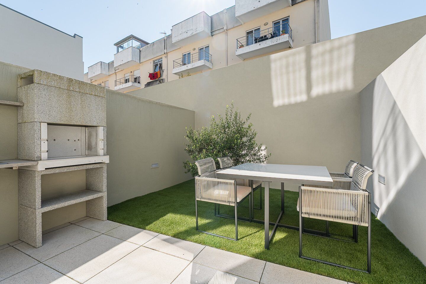 Une terrasse avec un barbecue, une table et des chaises, entourée d’une petite pelouse et d’un bâtiment en arrière-plan.