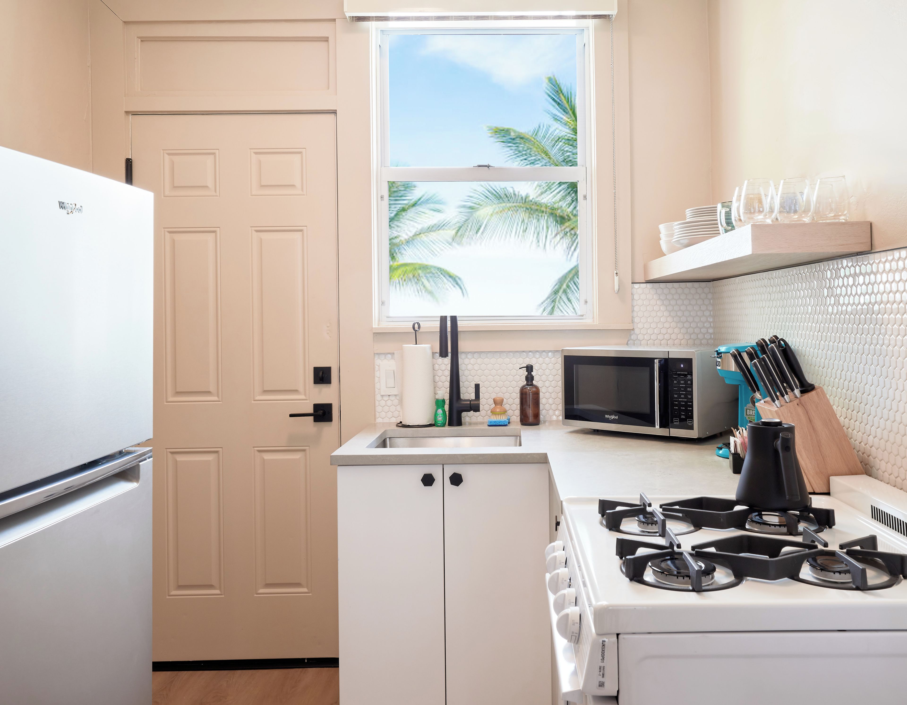 A compact kitchen with a refrigerator, microwave, stove, and a window with a view of palm trees.