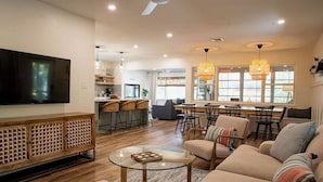 A spacious living room with a large TV, comfortable seating, a glass coffee table, and a kitchen area in the background. - Crabby Coconut-Mid-Island 3BR Perfect for Families! Sleeps 8 (Fort Myers Beach)