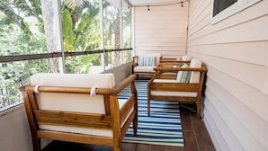 A wooden deck with two wooden chairs and a bench, a striped rug, and a view of greenery through large windows. - Crabby Coconut-Mid-Island 3BR Perfect for Families! Sleeps 8 (Fort Myers Beach)