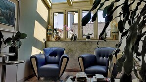 Ein Zimmer mit zwei blauen Ledersesseln, einem kleinen Tisch mit Büchern, einem Fenster mit Pflanzen und einem gerahmten Bild an der Wand.