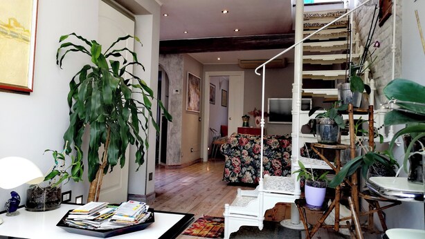 Ein geräumiger Wohnbereich mit Holzboden, einem weißen Sessel, einem Couchtisch mit Papieren, einer großen Grünpflanze und einer Treppe mit Holzwand.