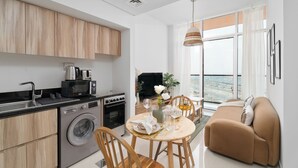 A modern kitchen with a washing machine, microwave, and coffee maker. There is a dining area with a table set for two, and a sofa in the living room.