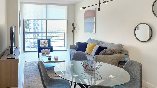 Una sala moderna con una mesa de comedor de cristal, sillas grises, un sofá con cojines, un televisor sobre un mueble bajo y un balcón con vistas.