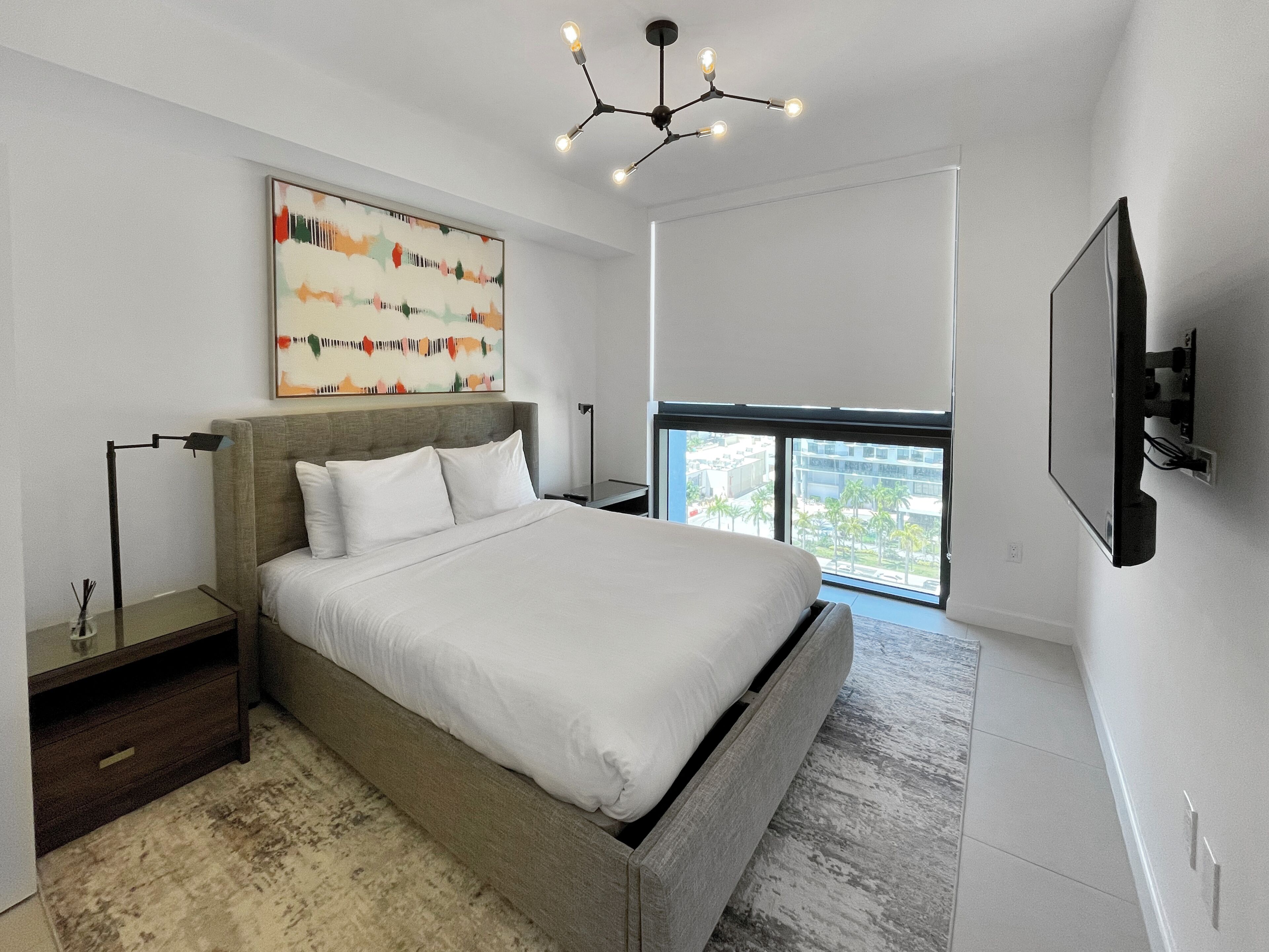 A modern bedroom with a large bed, a ceiling light fixture, a painting, and a TV mounted on the wall.