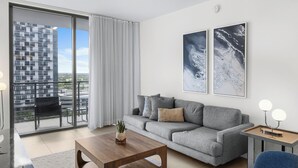 Een moderne woonkamer met een grijze bank, een houten salontafel en uitzicht op een wolkenkrabber door een schuifglazen deur.