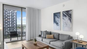 A modern living room with a grey sofa, a wooden coffee table, and a view of a high-rise building through a sliding glass door.