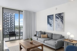 Un salon moderne avec un canapé gris, une table basse en bois et une vue sur un immeuble de grande hauteur, visible à travers une porte-fenêtre coulissante.