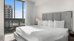 Een moderne hotelkamer met een groot bed, een nachtkastje, een lamp en een uitzicht op een stadslandschap door grote ramen.