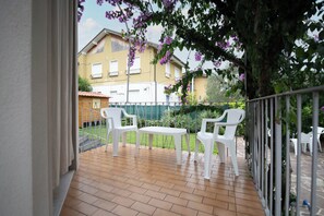 Un balcone con sedie di plastica bianca e un tavolino, affacciato su un giardino e degli edifici.