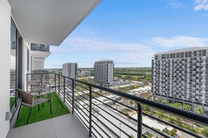 A balcony with a view of surrounding high-rise buildings and a grassy area with chairs.