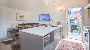 A modern kitchen with a central island, a dining area with a sofa and chairs, a mounted TV, and a view of a house with an American flag outside. - Downtown Dahl Haus: 2 bed/2 bath (Telluride)