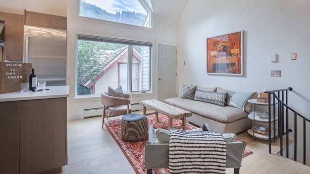 A modern living room with a sofa, a coffee table, and a dining area with a bench. There is a large window providing a view of the outdoors. - Downtown Dahl Haus: 2 bed/2 bath (Telluride)