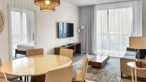 Una habitación de hotel moderna con una gran mesa redonda de comedor, asientos cómodos, un televisor de pantalla plana y ventanas amplias con cortinas traslúcidas.