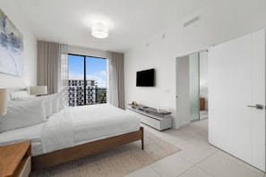 A modern hotel room with a large bed, a flat-screen TV, a balcony with a view of buildings, and a white door.