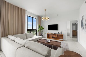 A modern living room with a sectional sofa, a coffee table, a TV, and a plant. - Elegant Doral Living: 3BR, 3 Baths (Doral)