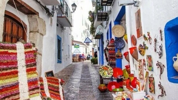 Une rue étroite bordée de bâtiments blancs, de balcons ornés de plantes et de divers objets colorés mis en vente.
