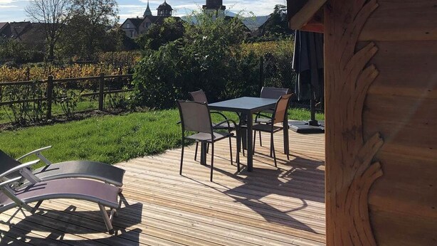 Une terrasse en bois avec des meubles d’extérieur, un vignoble en arrière-plan et le clocher d’une église.